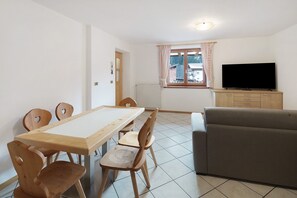 Un coin repas avec une table et des chaises en bois, un canapé gris, un téléviseur posé sur un meuble en bois et une fenêtre avec des rideaux.