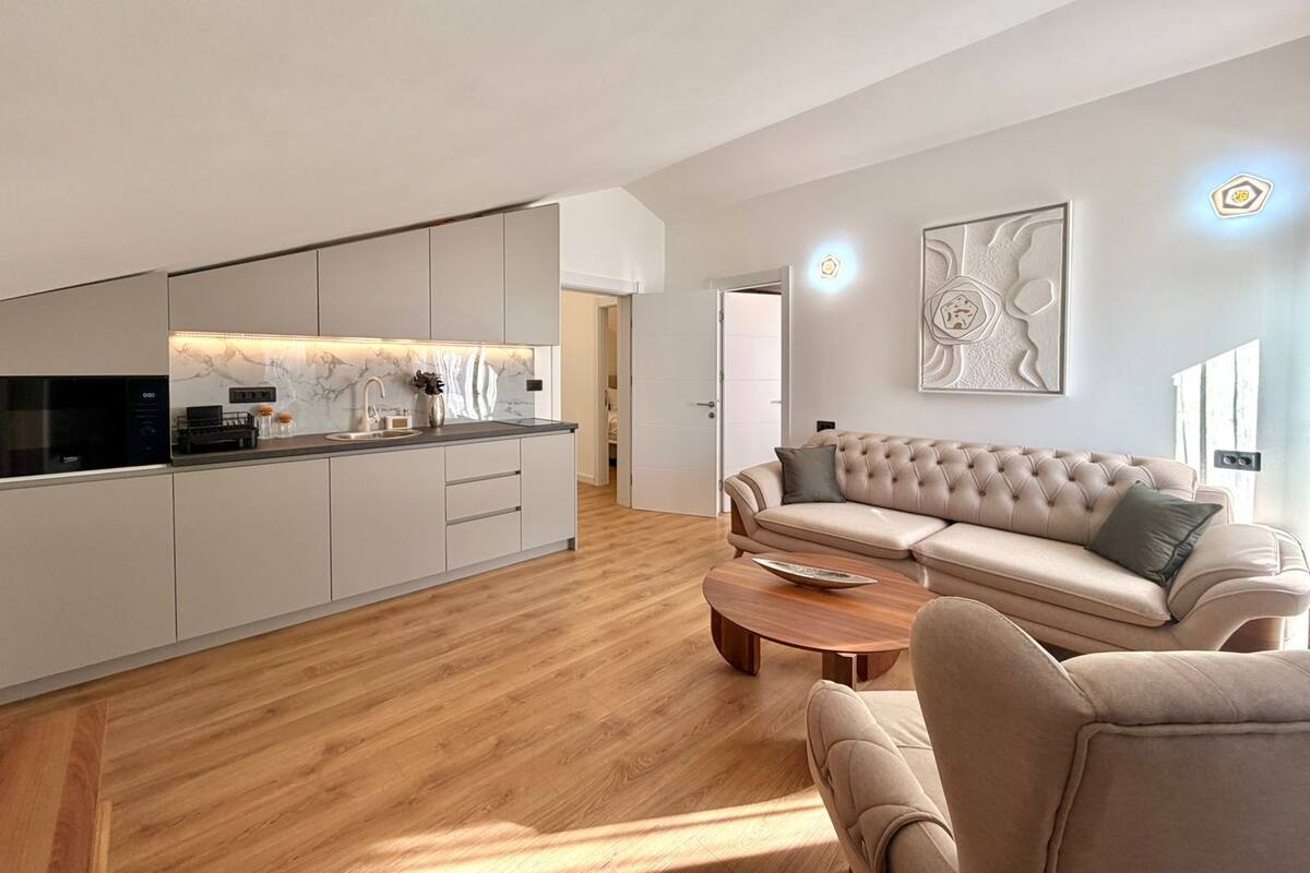 A modern living room with a sofa and coffee table, a kitchen with a marble backsplash, and a large abstract painting on the wall.
