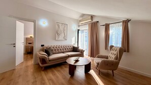 A modern living room with a beige sofa, a round coffee table, and a matching armchair. There is a door leading to a smaller room with a vanity and a mirror.