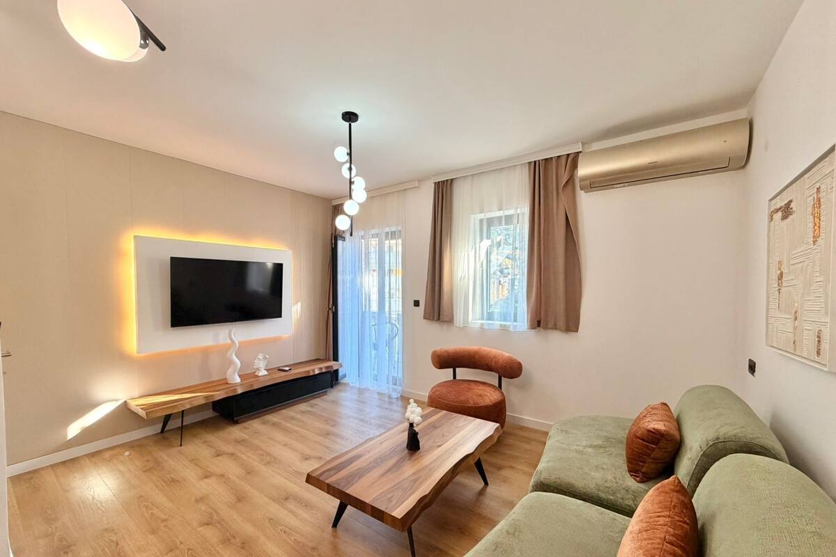 A modern living room with a flat-screen TV mounted on the wall, a wooden coffee table, a green sofa with cushions, and a pendant light.