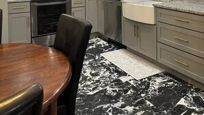 Una cocina moderna con armarios grises, un microondas de acero inoxidable, una estufa y un lavamanos. Hay una mesa de comedor con sillas y una alfombra estampada en blanco y negro.