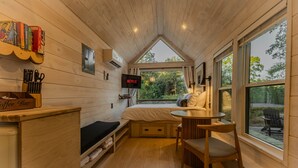 A cozy cabin interior with a bed, a coffee bar, a TV, and large windows offering a view of the outdoors. - Zen Den Cabin (Houston) (Coldspring)
