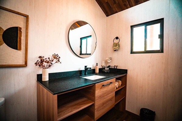 A modern bathroom with a wooden vanity, a round mirror, a window, and a framed artwork on the wall.