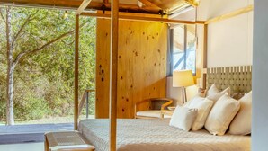 Een slaapkamer met een groot bed, een houten hoofdbord, een nachtkastje met een lamp en uitzicht op bomen.