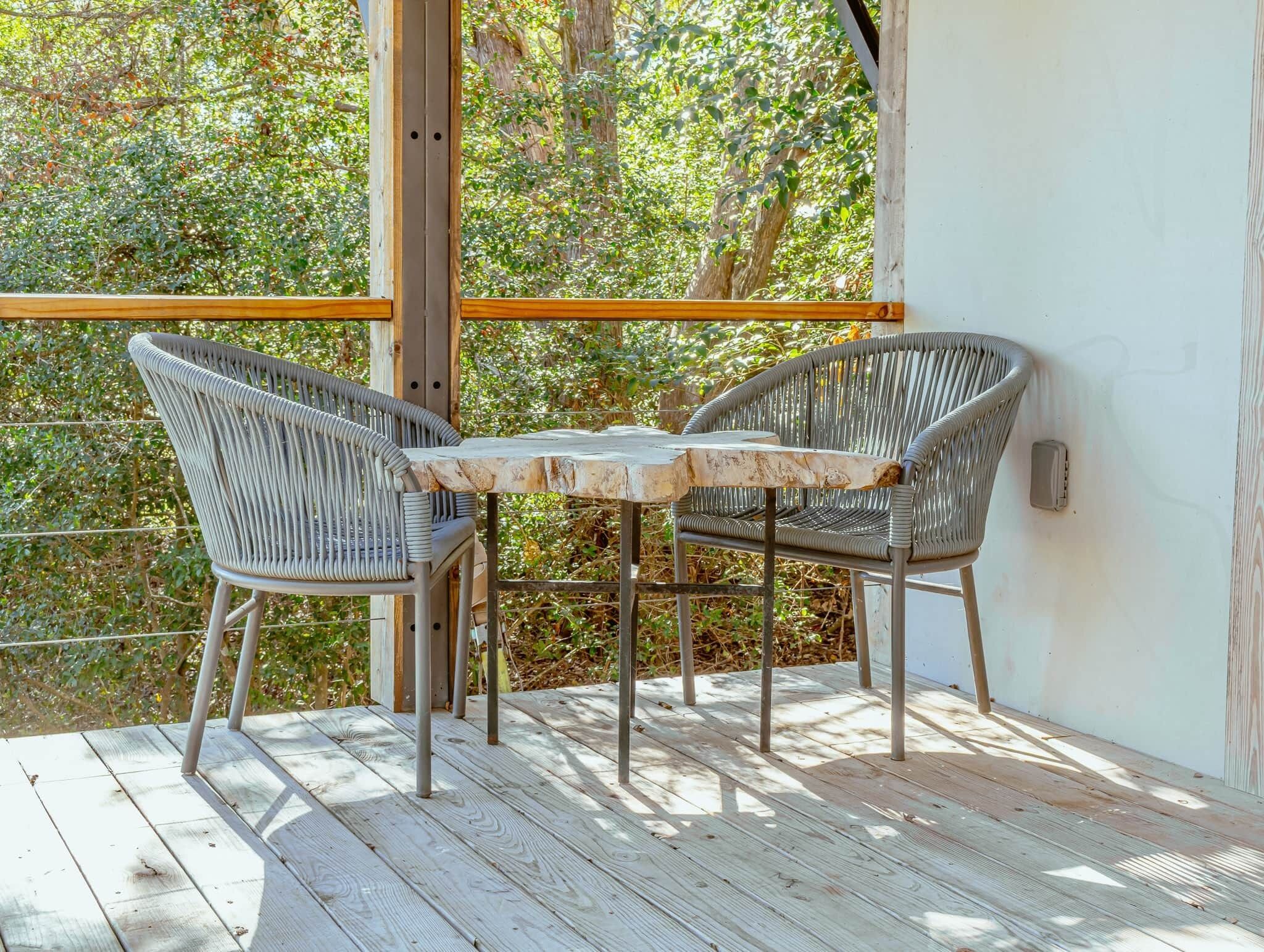 En træterrasse med to fletstole og et lille bord, omgivet af et trægelænder og en hvid væg.