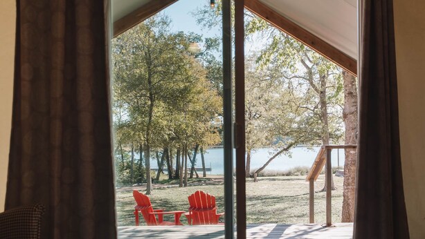 Ein Schlafzimmer mit einem großen Bett, einer Glasschiebetür, die zu einer Terrasse im Freien mit roten Stühlen und Blick auf Bäume und Wasser führt.