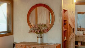 Un mobiletto in legno con un vaso in metallo pieno di fiori, uno specchio rotondo e una scaffalatura in legno con un lavandino.