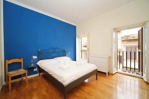 Un dormitorio con cama, una silla, una mesita de noche y una ventana con vistas a edificios.
