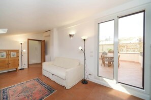 Ein Wohnzimmer mit einem weißen Sofa, einer Stehlampe, einem gemusterten Teppich, einer Holzkabine und einer Glastür, die zu einer Außenpatio führt.