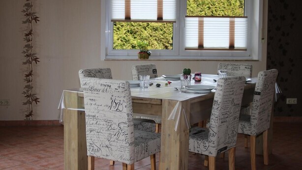 Een eetgedeelte met een tafel gedekt voor vier personen, omringd door stoelen met handgeschreven aantekeningen, een raam met zonwering en een bloempot op de vensterbank.