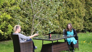 Twee personen zitten aan een tafel met glazen blad buiten, omgeven door groen.