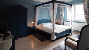 A bedroom with a four-poster bed, a nightstand, a chair, a wardrobe, and a window with curtains.