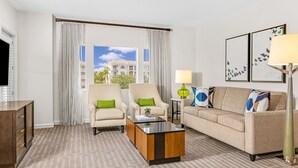 A hotel room with a large window, two armchairs, a sofa, a coffee table, a lamp, and artwork on the wall.