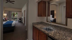 A hotel room with a bed, a desk, a chair, a small table, a mirror, a sink, and a ceiling fan.