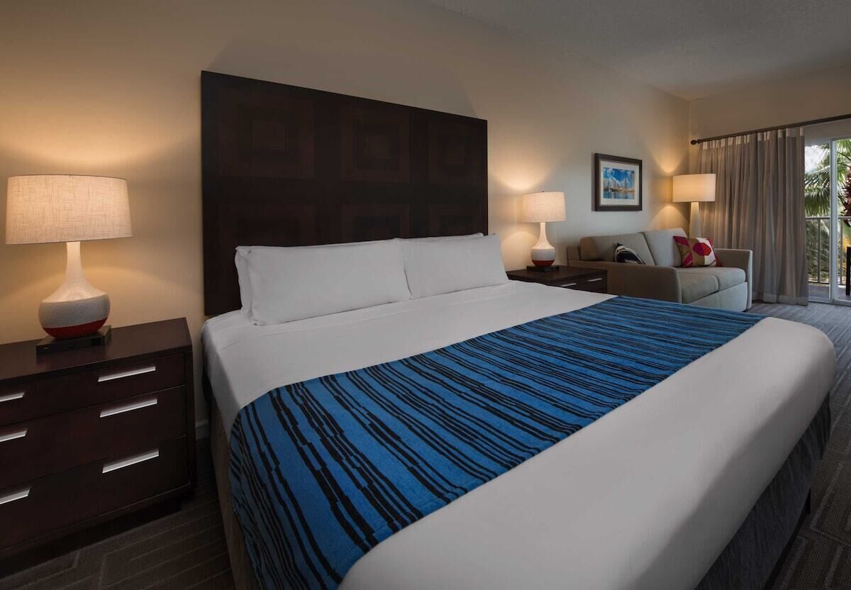 Een hotelkamer met een groot bed, twee nachtkastjes met lampen, een bank, een raam met gordijnen en een ingelijst schilderij aan de muur.