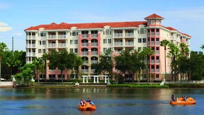 A multi-story hotel with balconies and a red-tiled roof, situated by a calm body of water with paddle boats.