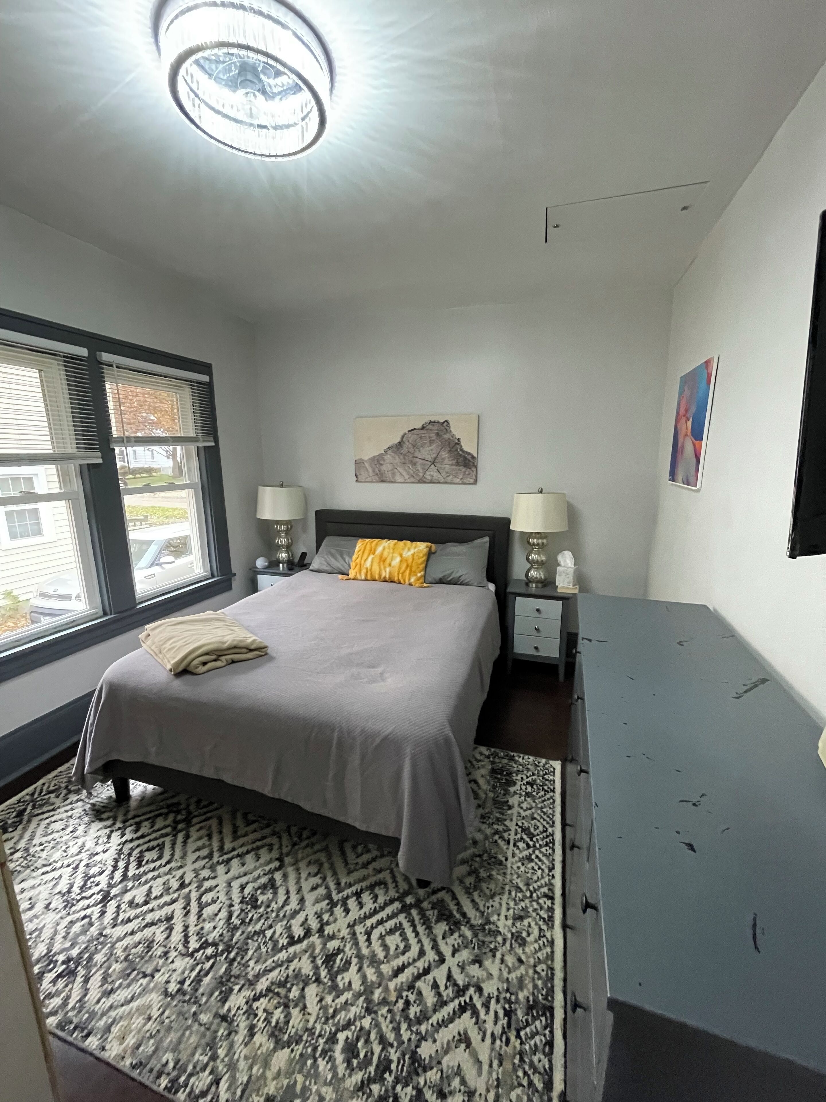 A bedroom with a bed, bedside lamps, a rug, a desk, and a window with blinds.