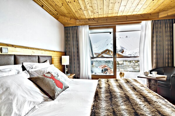 A hotel room with a wooden ceiling, a bed with white linens, a fur throw, a view of a mountain, and a small table with a cup and saucer. - Hôtel Saint Martin (Les Belleville)