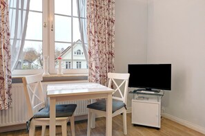 A room with a dining table, chairs, a television on a stand, and a window with curtains.