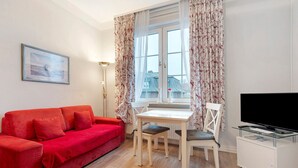A room with a red sofa, a dining table with chairs, a TV on a white cabinet, and a window with floral curtains. - Apartment 'Anker' with Shared Terrace and Wi-Fi (Cuxhaven)