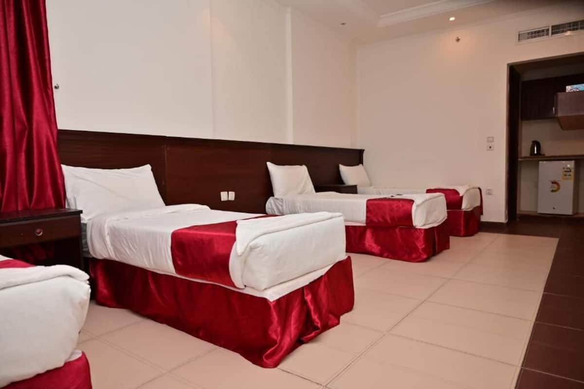 Une chambre d’hôtel avec trois lits, chacun doté de draps blancs et de couvertures rouges, d’une tête de lit en bois et d’un petit coin cuisine.