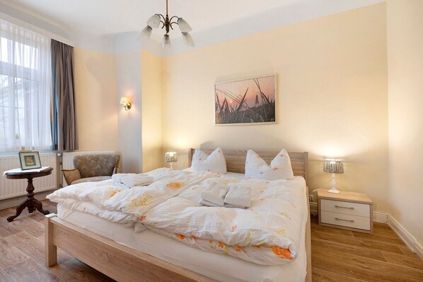 A neatly made bed with a floral duvet, a bedside table with a lamp, a chair, and a picture on the wall.