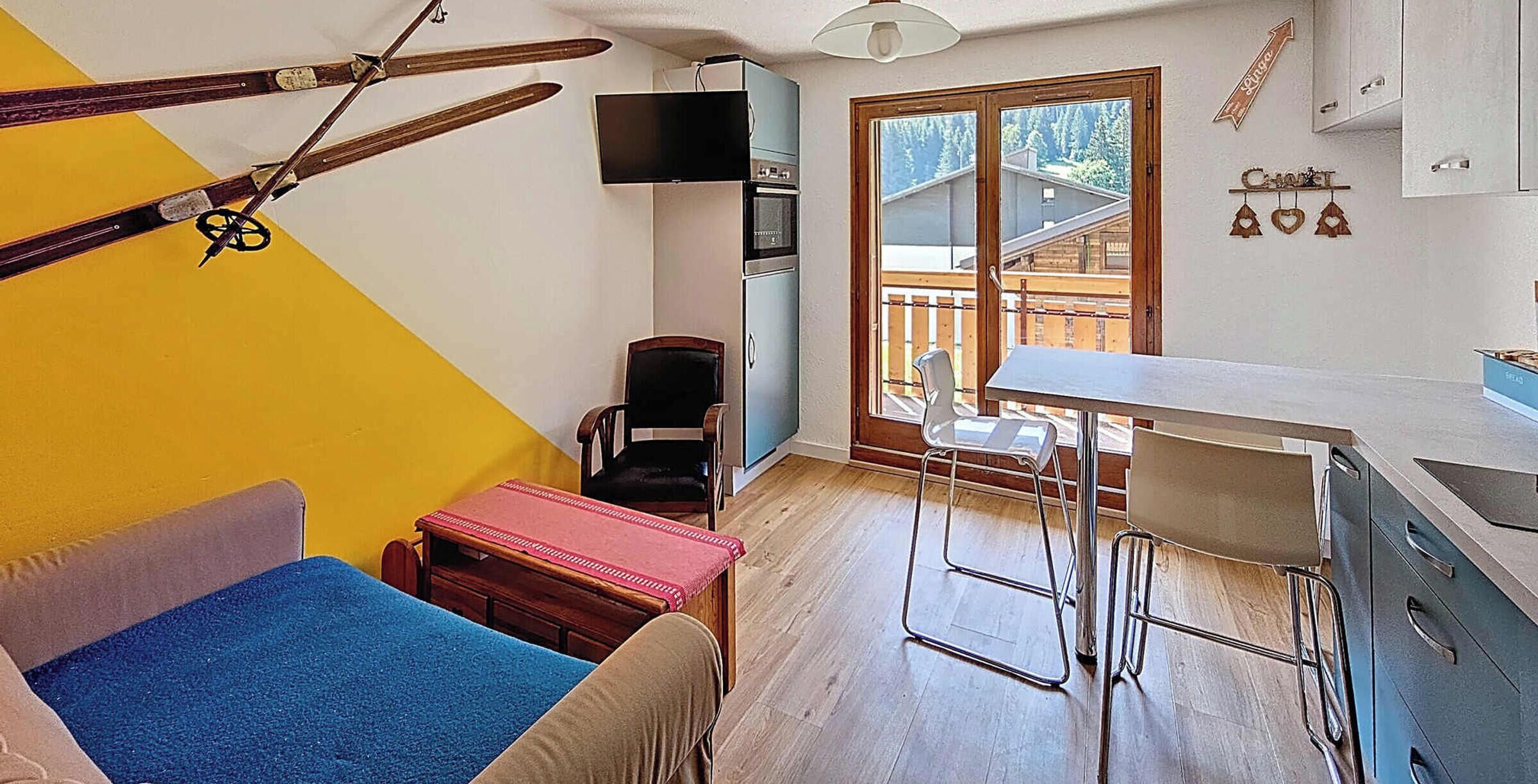 Une pièce de taille modeste comprenant un canapé, une petite table, un tabouret de bar, un téléviseur, une porte en bois donnant sur un balcon et un porte-skis.