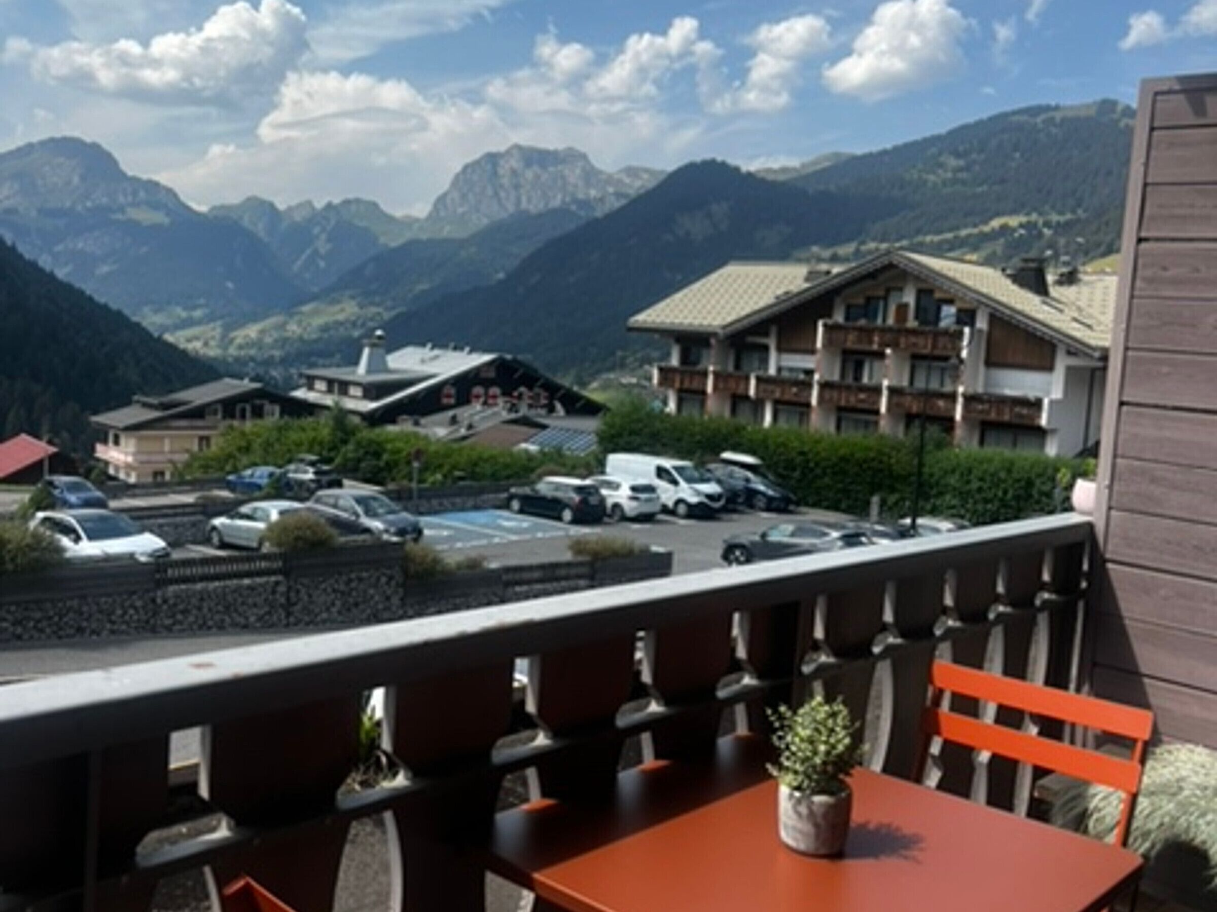 A mountain view with a dining area featuring a table and chairs, a parking lot with several cars, and a building with multiple floors in the background.