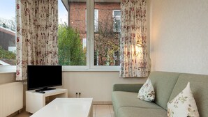 A living room with a TV on a stand, a white table, a green sofa with floral cushions, and a window with curtains.