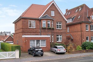 Une maison à deux étages, construite en briques et surmontée d’un toit recouvert de tuiles rouges, avec une voiture garée dans l’allée et une clôture blanche.
