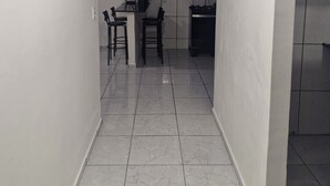 A narrow hallway with white walls and a tiled floor leading to a kitchen area with a dining table and chairs. - Cozy place in Perdeira (Belém)