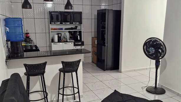 A modern kitchen with a built-in oven, microwave, and refrigerator. There are two black bar stools and a black sofa with a cushion. A blue water dispenser is on the left, and a standing fan is on the right. - Cozy place in Perdeira (Belém)