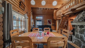 Interior acogedor de una cabaña de troncos, con comedor que incluye una mesa de madera, sillas y una chimenea de piedra.
