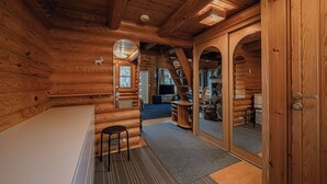 Interior de una cabaña de troncos con un banco de madera, una mesita y una puerta corrediza que da a una zona con chimenea.