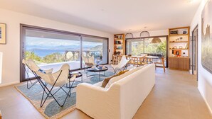Ein modernes Wohnzimmer mit einem großen Schiebeglastür, die zu einem Balkon mit schöner Aussicht führt. Es gibt ein weißes Sofa, zwei weiße Sessel und eine Essecke mit Tisch und Stühlen.