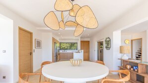 A modern kitchen with white cabinets, a central island, and a dining area with a round table and wooden chairs. - VILLA MAGUY – 3 Bedrooms, Swimming Pool, Quiet and Secluded Location (PIETROSELLA)