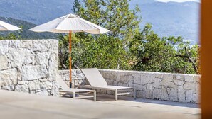 A sunlit outdoor seating area with a stone wall and a view of mountains. - VILLA MAGUY – 3 Bedrooms, Swimming Pool, Quiet and Secluded Location (PIETROSELLA)