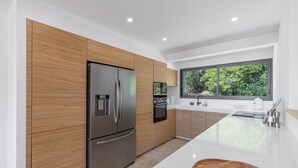 A modern kitchen with wooden cabinets, a stainless steel refrigerator, and a large window offering a view of greenery. - VILLA MAGUY – 3 Bedrooms, Swimming Pool, Quiet and Secluded Location (PIETROSELLA)
