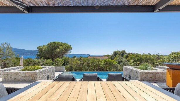 A wooden deck with a pool, stone walls, and outdoor seating. - VILLA MAGUY – 3 Bedrooms, Swimming Pool, Quiet and Secluded Location (PIETROSELLA)