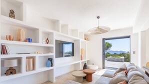 A modern living room with a large TV, white shelving, a sofa, and a view of the outdoors. - VILLA MAGUY – 3 Bedrooms, Swimming Pool, Quiet and Secluded Location (PIETROSELLA)