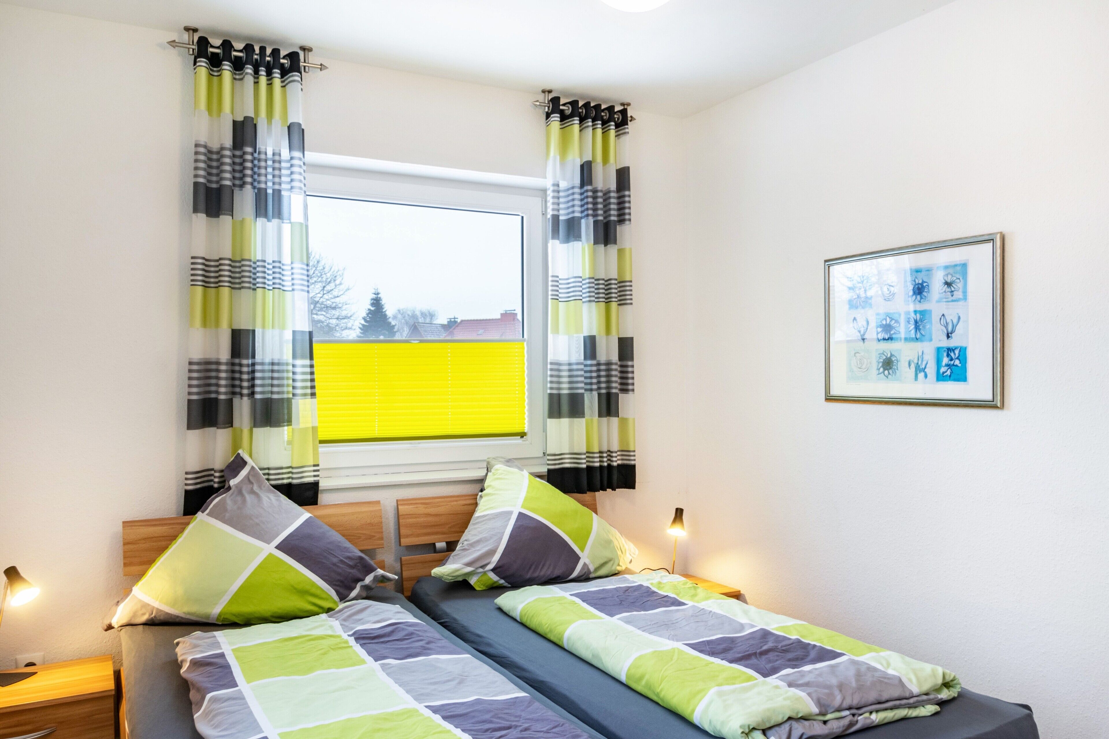 Una habitación con dos camas, una ventana con cortinas, un cuadro enmarcado en la pared y una mesita de noche con una lámpara.