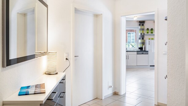 A hallway with a mirror, a lamp, and a book on a counter. There is a door leading to a kitchen area. - Apartment 'Ferienwohnung' with Balcony and Wi-Fi (Horumersiel-Schillig)