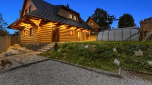 Hirsimökki, jossa on kalteva katto, kuisti ja taustalla kasvihuone.