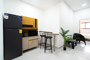 A compact kitchen with a black refrigerator, wooden cabinets, a microwave, and a small dining area with two black stools and a potted plant.