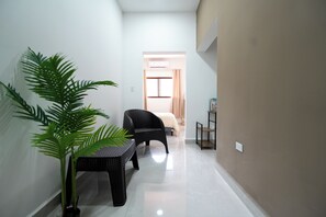 A narrow hallway with a black wicker chair, a small table with a potted plant, and a bed in the background.