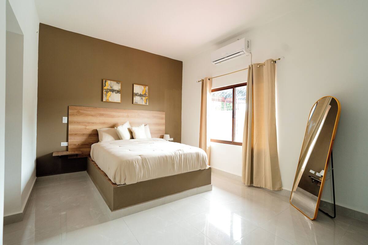 A modern bedroom with a large bed, a bedside table, a wall-mounted air conditioner, and a window with curtains.