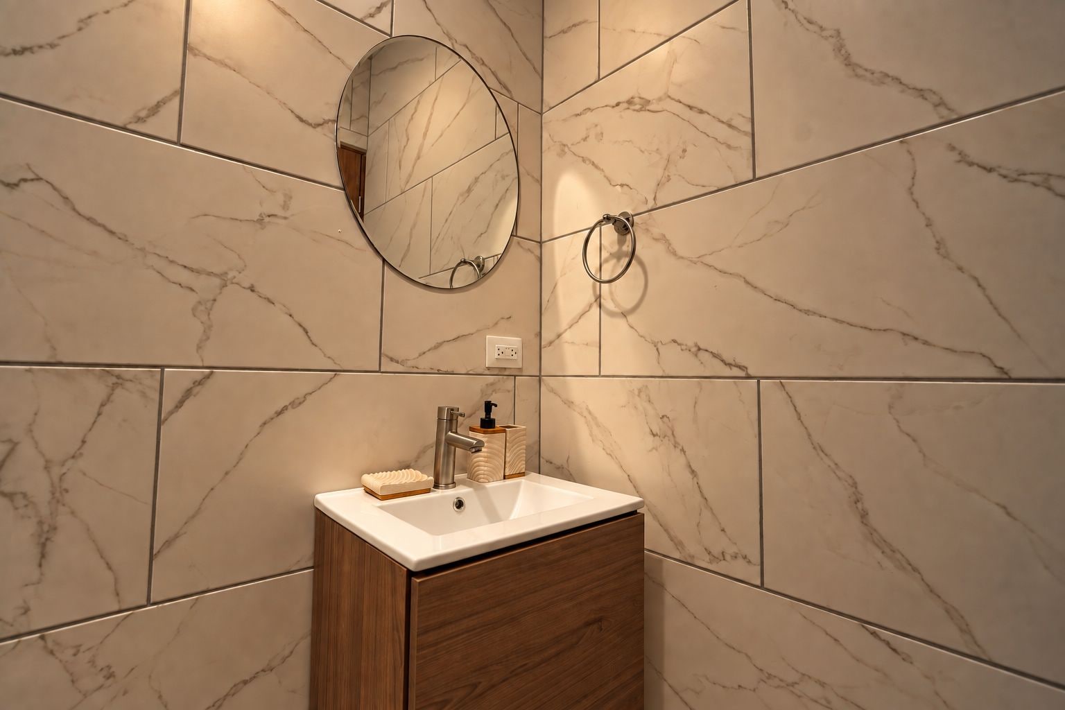 Une salle de bain avec un mur recouvert de carreaux de marbre, un meuble en bois, un lavabo blanc, un miroir et un porte-serviettes.