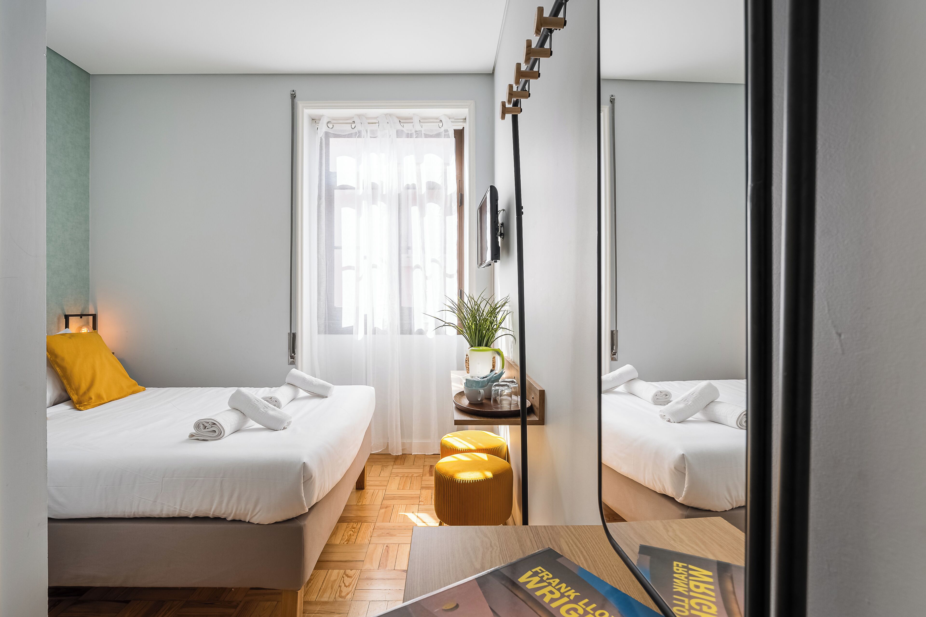 A modern hotel room with two beds, a small table with a plant, a mirror, and a window with curtains.