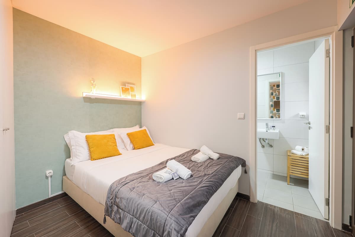 Una camera d'albergo con un letto, un comodino, uno specchio e un bagno visibile attraverso una porta aperta.