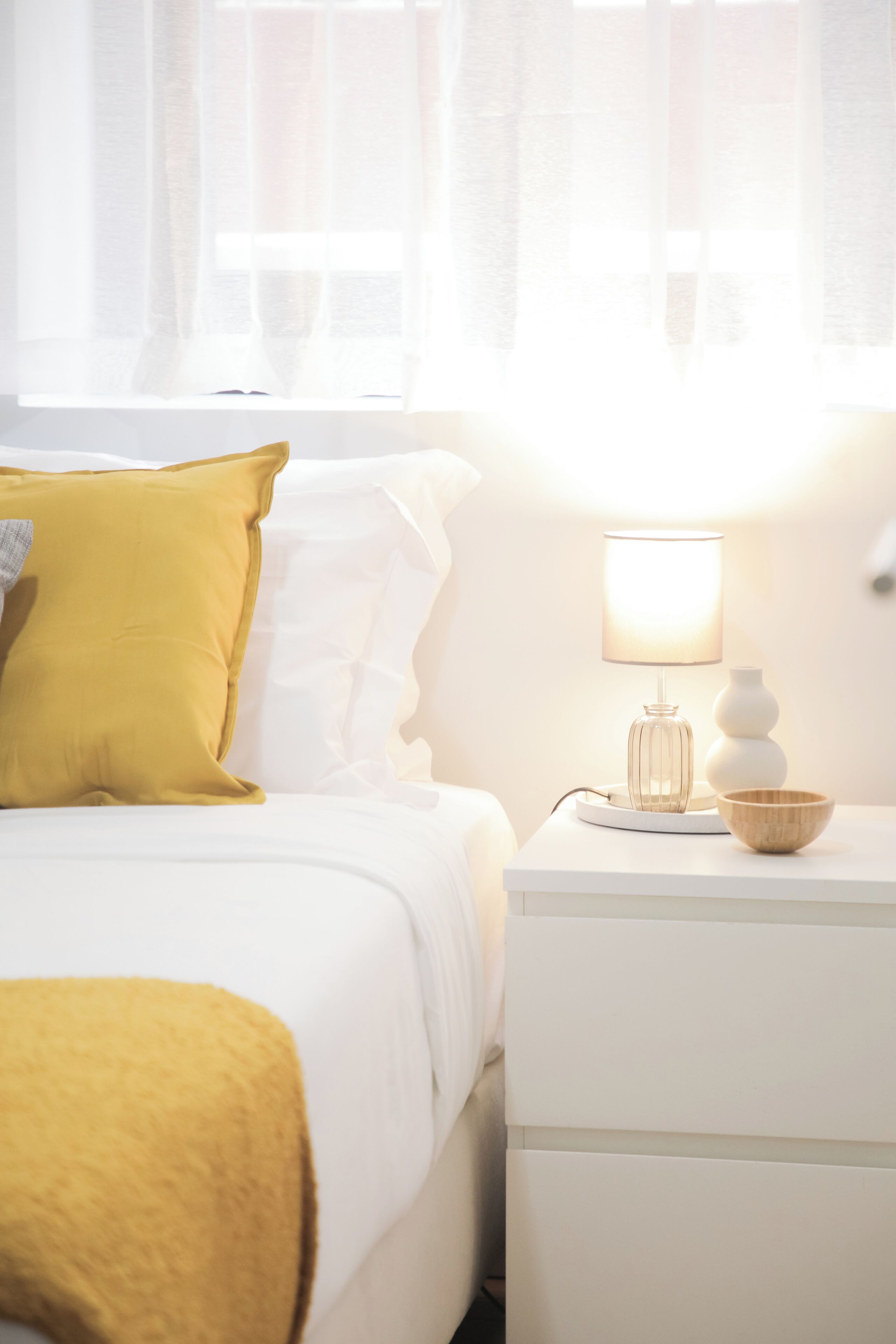 A neatly made bed with a yellow pillow, a white bedside table with a lamp, and a vase.